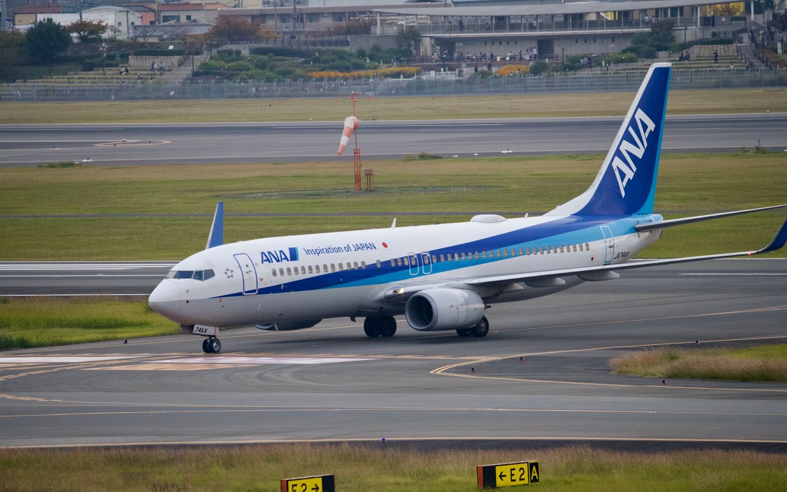 An ANA Boeing 737 taxiing across a grassy airfield, with a terraced viewing area behind