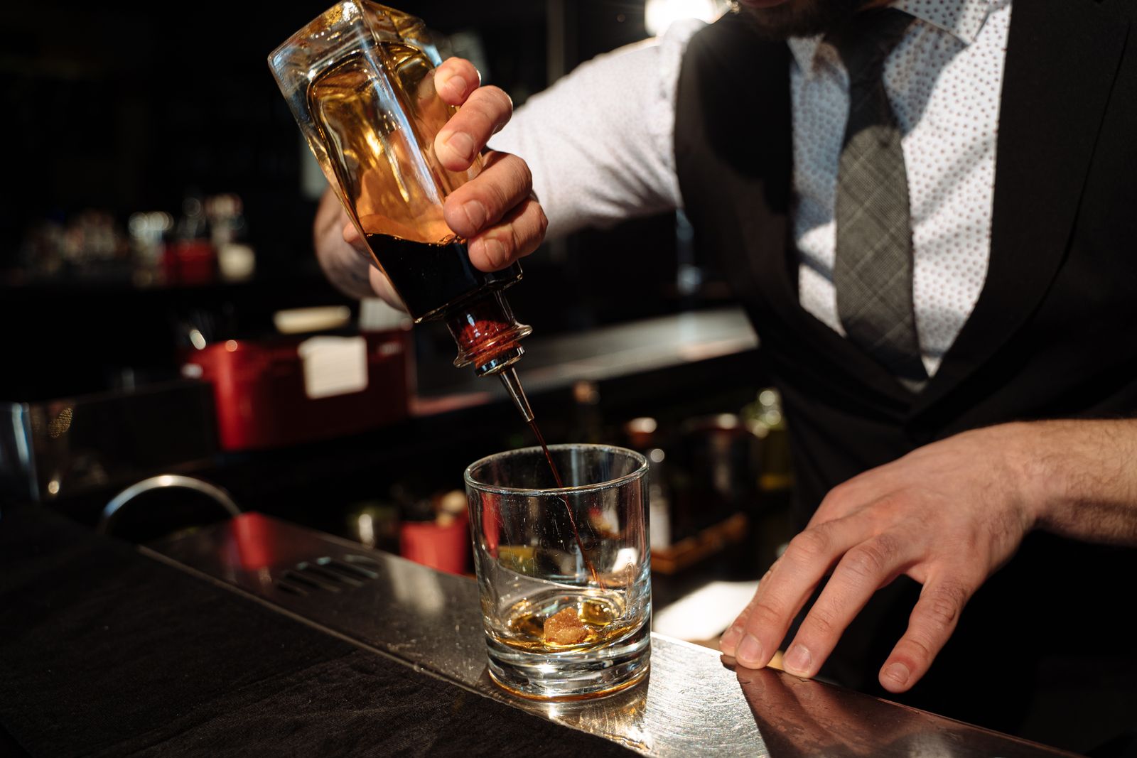 A bartender in a waistcoat and tie pouring whisky into a glass over a large ice cube