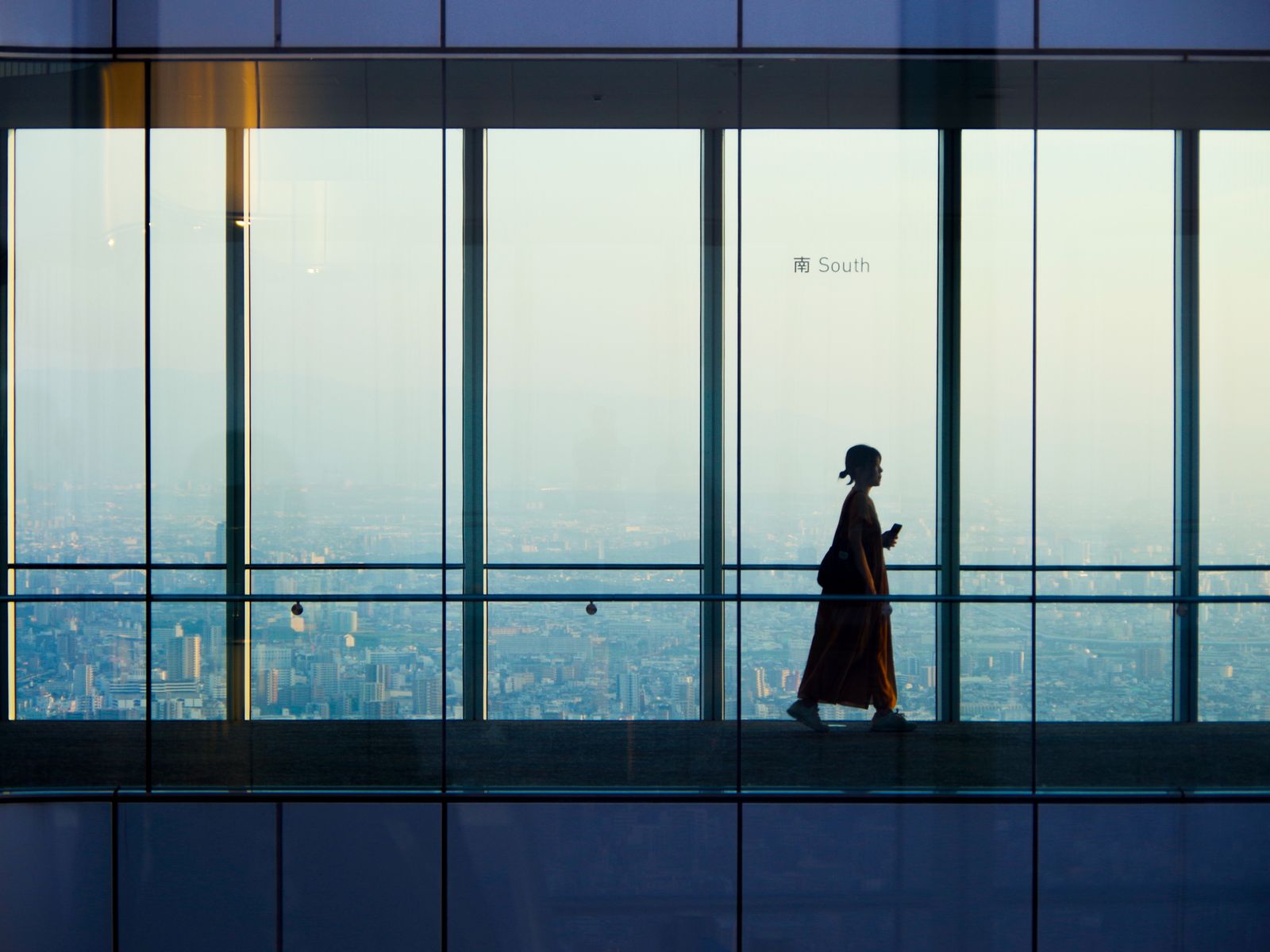 A visitor silhouetted against floor-to-ceiling observatory windows marked South, with Osaka spread flat to the horizon
