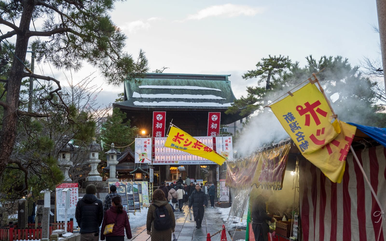 Visitors walking past steaming food stalls towards a temple gate hung with hatsumode banners in winter