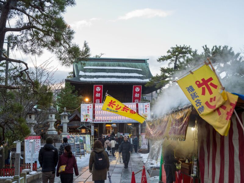 Visitors walking past steaming food stalls towards a temple gate hung with hatsumode banners in winter
