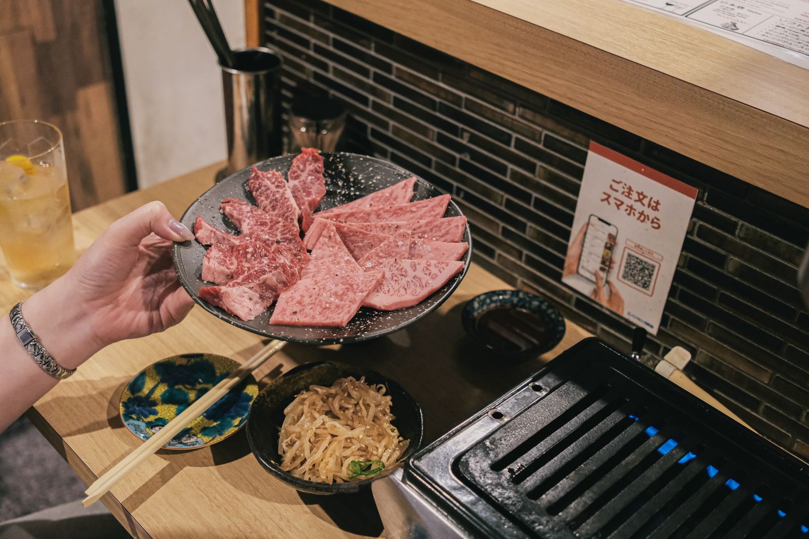 A diner holding a plate of marbled beef at a one-person yakiniku counter with its own small gas grill