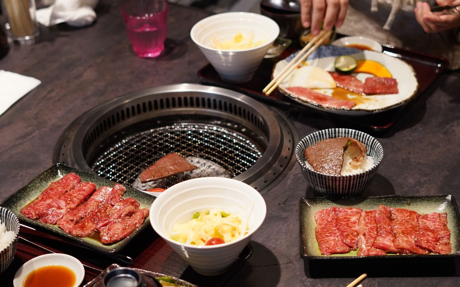 A yakiniku table with plates of marbled beef, rice bowls and salad set around a round built-in grill