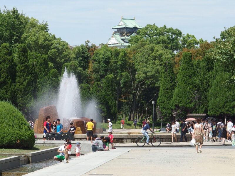 Adults and children gather beside a fountain in Osaka Castle Park, with the castle keep rising above green trees.