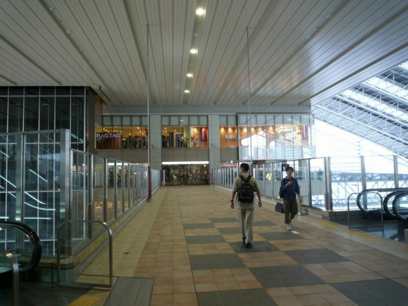A backpack-wearing pedestrian crosses a covered walkway at Osaka Station City, beside glass railings, escalators and another passerby.