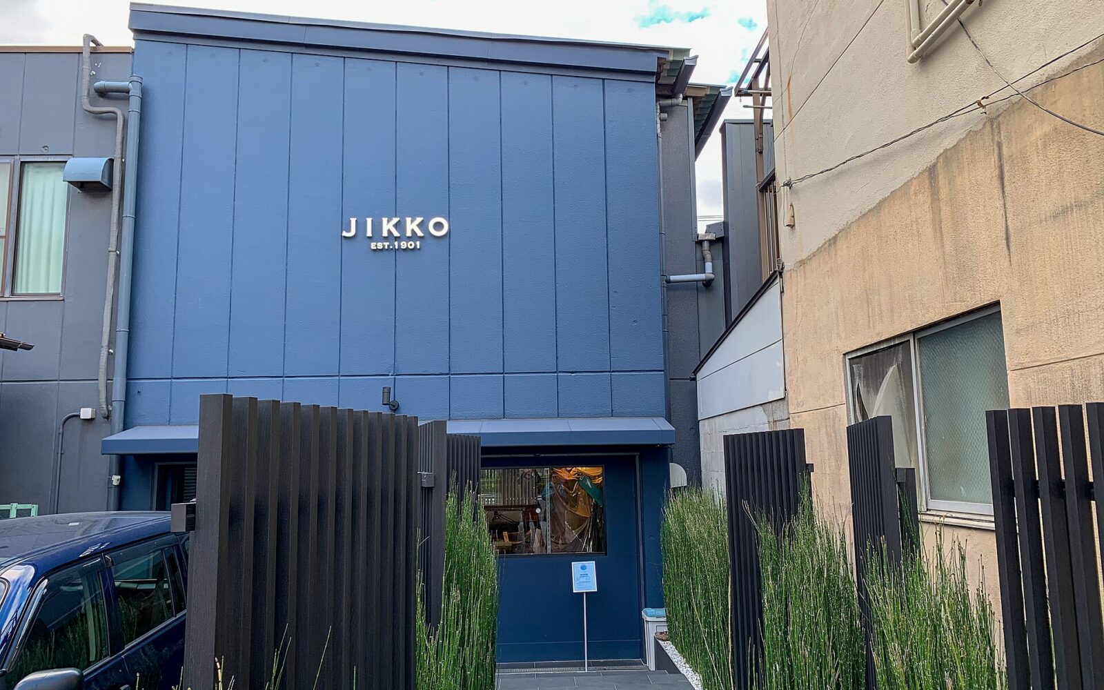 Blue facade of the Jikko knife shop in Sakai, with a recessed entrance between tall green plants.