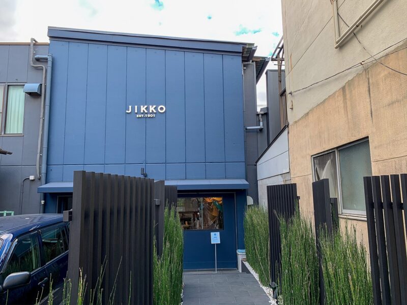 Blue facade of the Jikko knife shop in Sakai, with a recessed entrance between tall green plants.