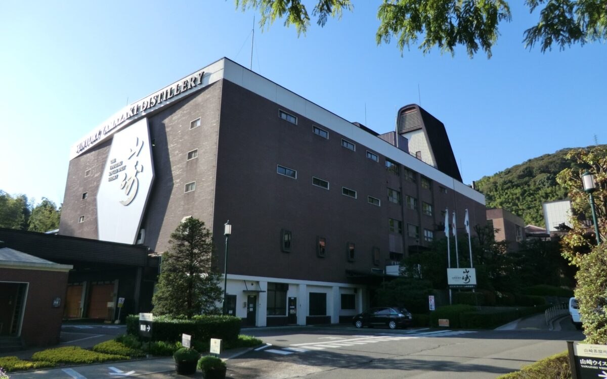 The brick Yamazaki Distillery building in Shimamoto, Osaka Prefecture, beside trees and a sunlit driveway.
