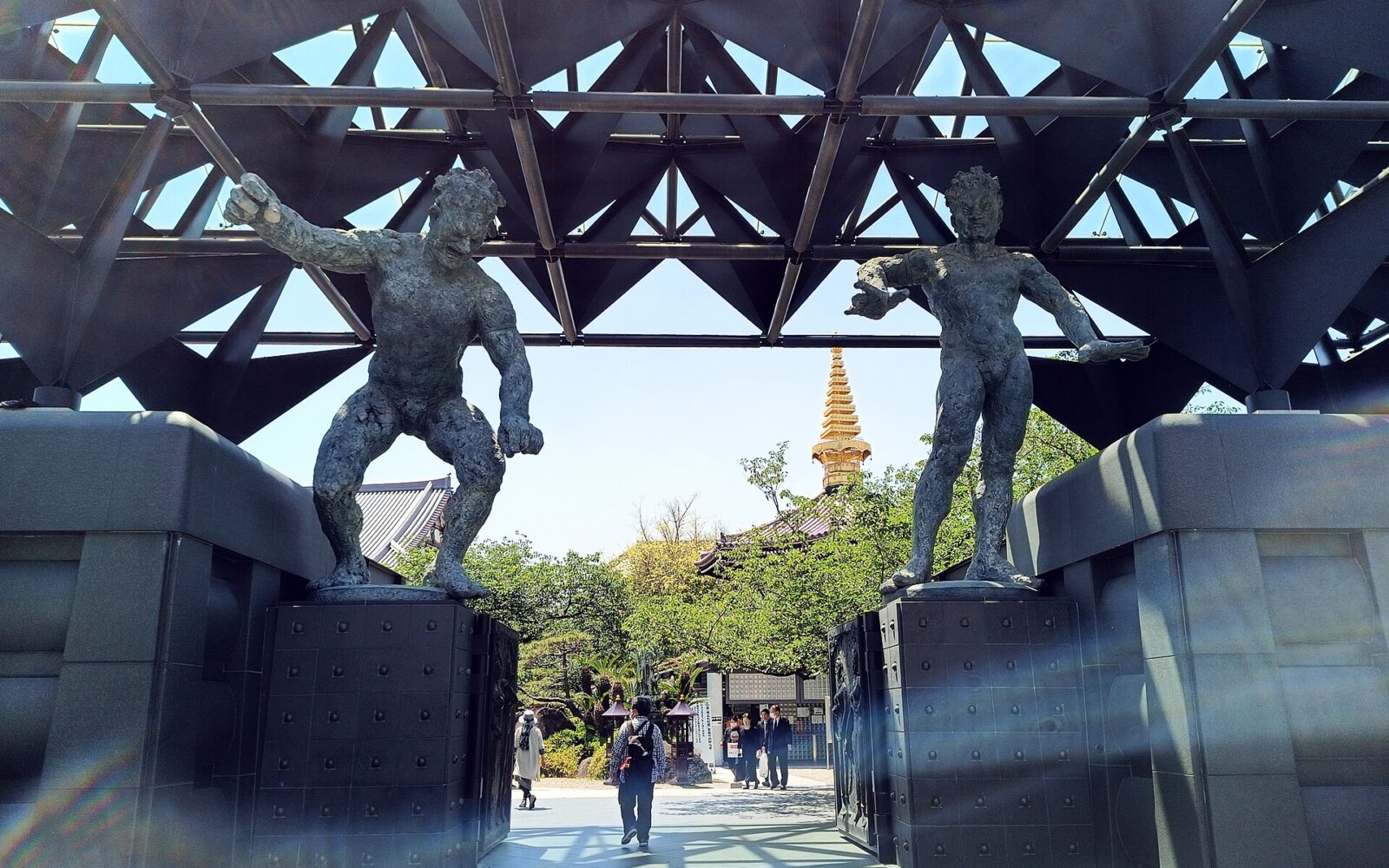 Bronze guardian figures flank Isshinji’s entrance in Osaka beneath a geometric steel roof, with visitors walking into the temple grounds.