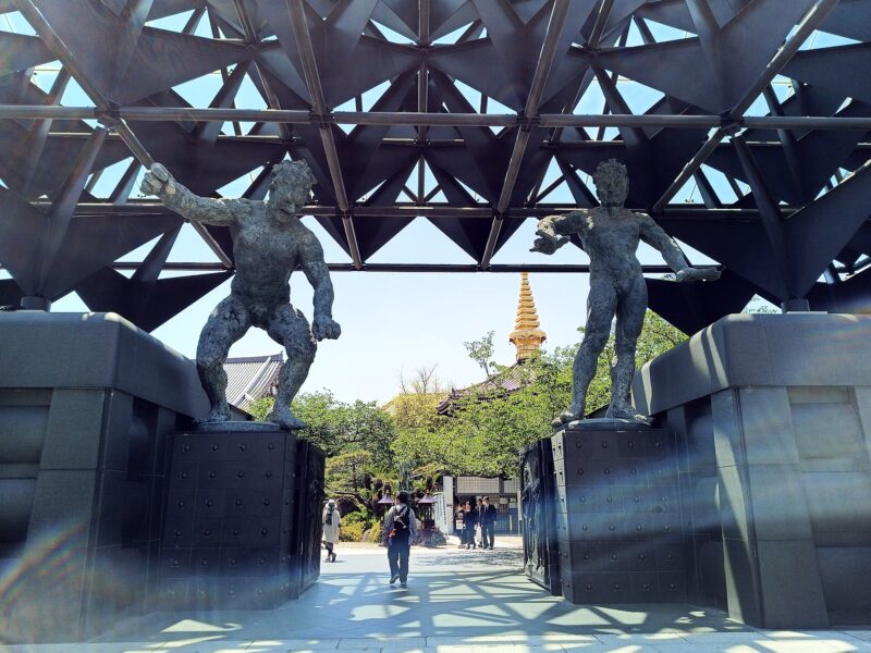 Bronze guardian figures flank Isshinji’s entrance in Osaka beneath a geometric steel roof, with visitors walking into the temple grounds.
