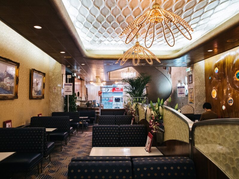 Cafe American in Osaka has upholstered booths, wood-paneled walls, ornate chandeliers and a curved staircase at the far end.