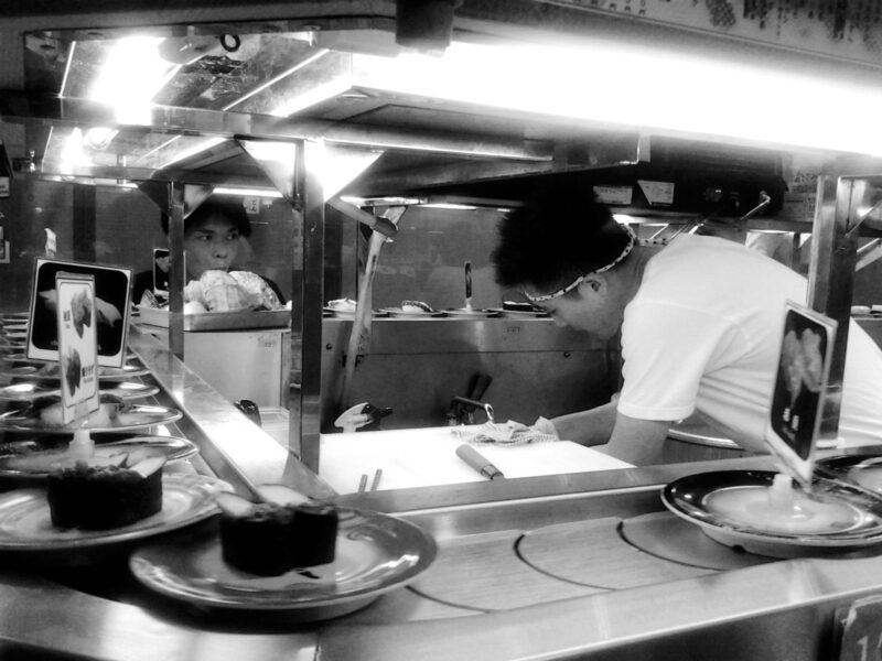 Chefs prepare sushi behind a conveyor belt carrying small plates at a sushi counter in Osaka.