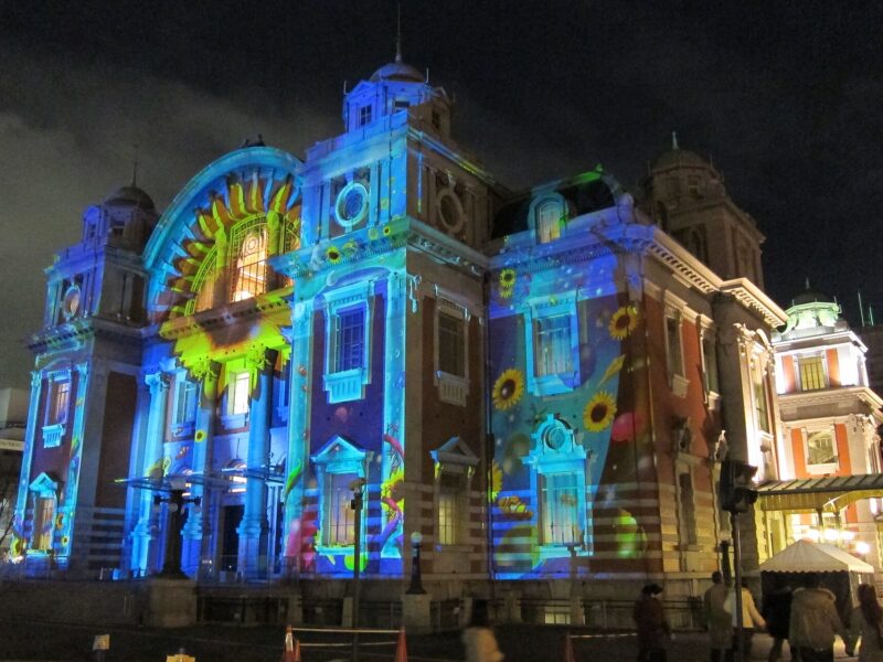 Colorful floral projections illuminate Osaka Central Public Hall at night, with visitors beside the building.
