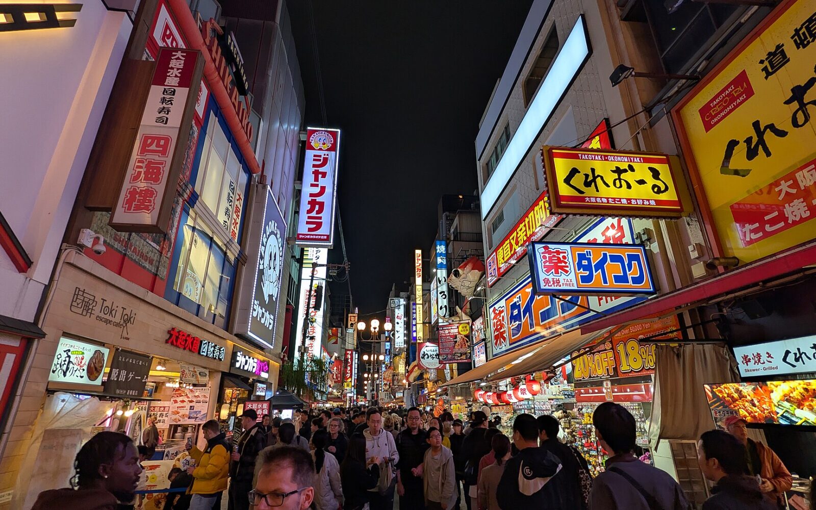 Crowds walk beneath bright restaurant signs along Dotonbori in Osaka after dark, with food counters lining the street.