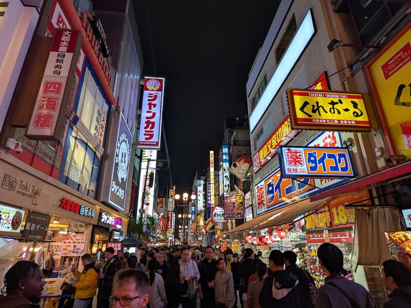 Crowds walk beneath bright restaurant signs along Dotonbori in Osaka after dark, with food counters lining the street.