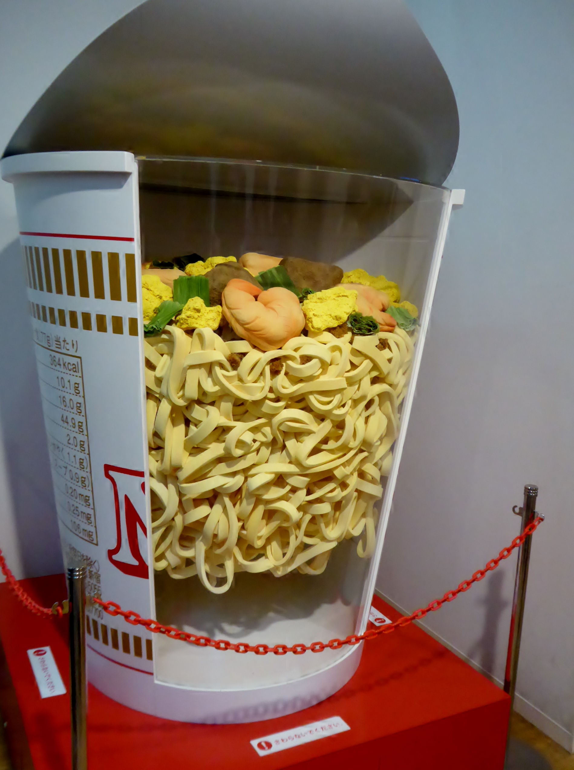 A giant cutaway model of a CUP NOODLE cup showing layers of noodles with shrimp, egg and vegetable pieces on top, on a red plinth behind a rope