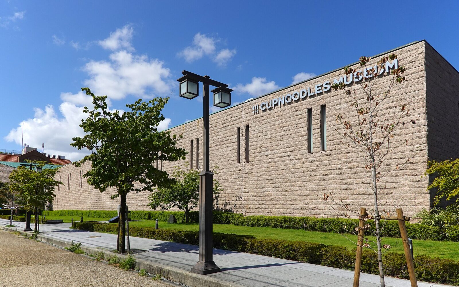 The long stone-clad wall of the CUPNOODLES MUSEUM in Ikeda, with its name in large letters, a lawn, young trees and a street lamp under a blue sky
