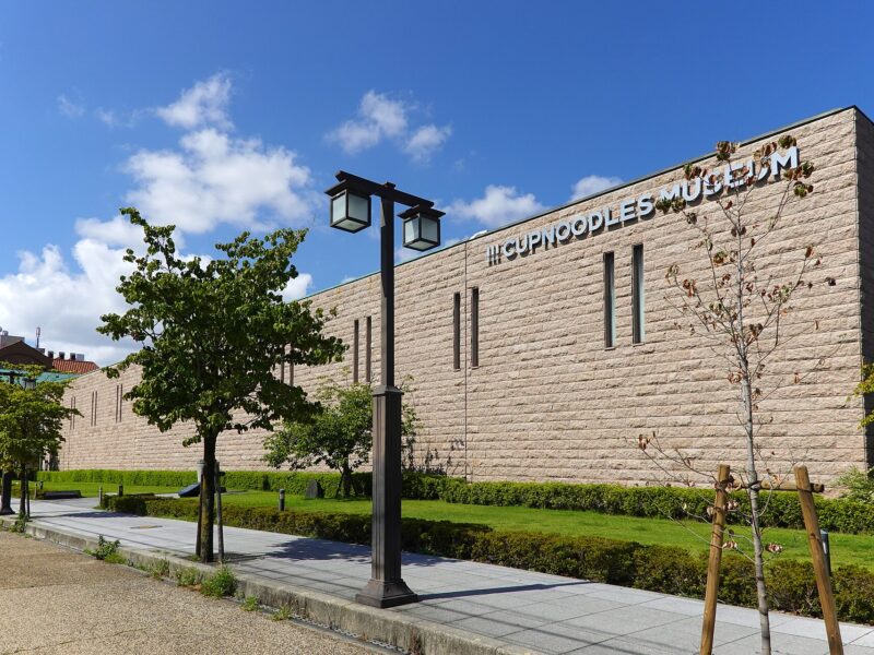 The long stone-clad wall of the CUPNOODLES MUSEUM in Ikeda, with its name in large letters, a lawn, young trees and a street lamp under a blue sky