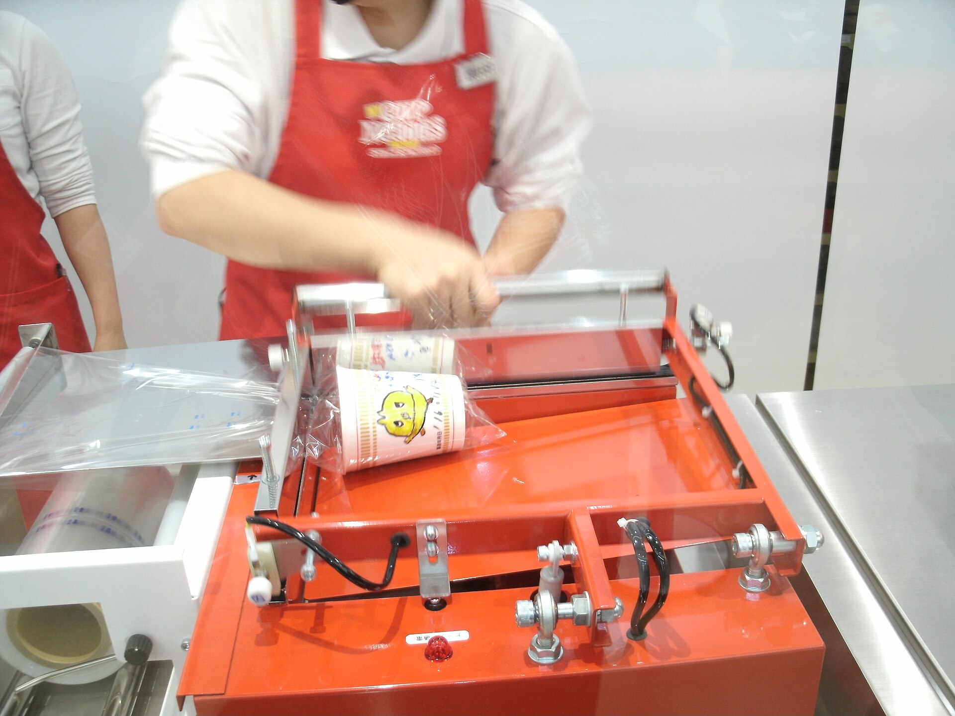 A staff member in a red apron operating a red sealing machine that holds a decorated cup of noodles inside a clear plastic wrapper