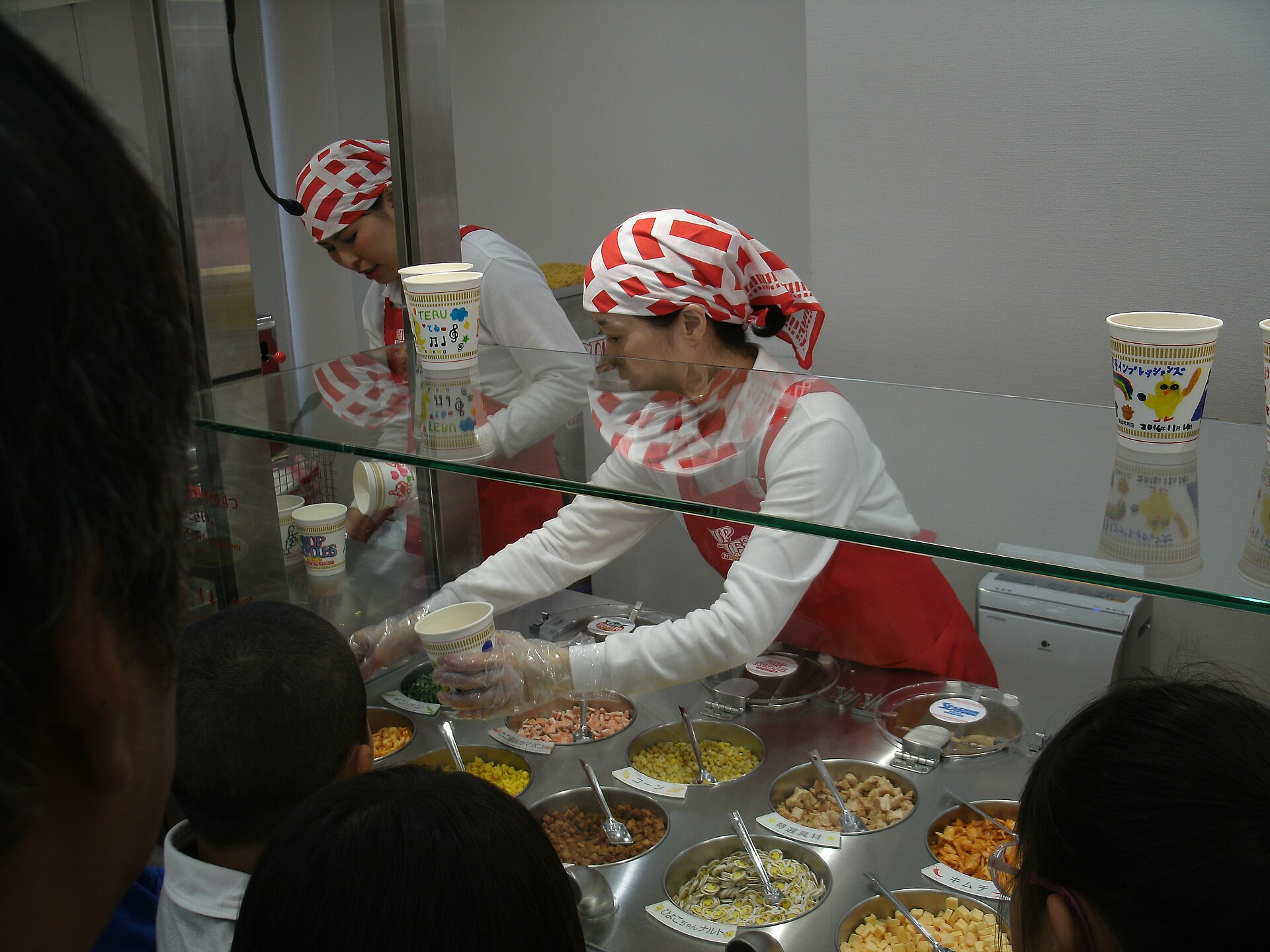 Museum staff in red and white headscarves and aprons filling decorated cups with dried noodle toppings from steel trays behind a glass counter