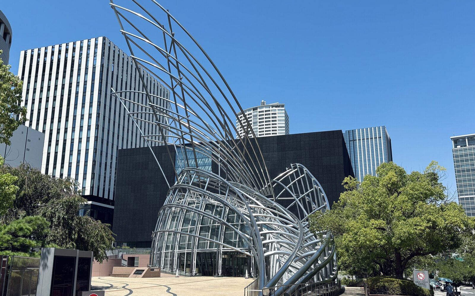 Curving silver tubes rise above the glass entrance of the National Museum of Art, Osaka, beneath a blue sky.