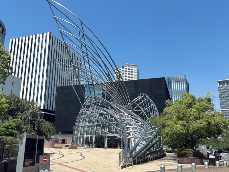 Curving silver tubes rise above the glass entrance of the National Museum of Art, Osaka, beneath a blue sky.