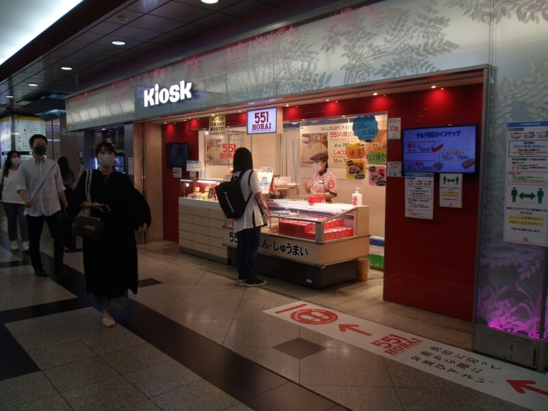 A customer stands at the 551 Horai counter in Osaka Station, where red takeaway boxes fill the glass display.