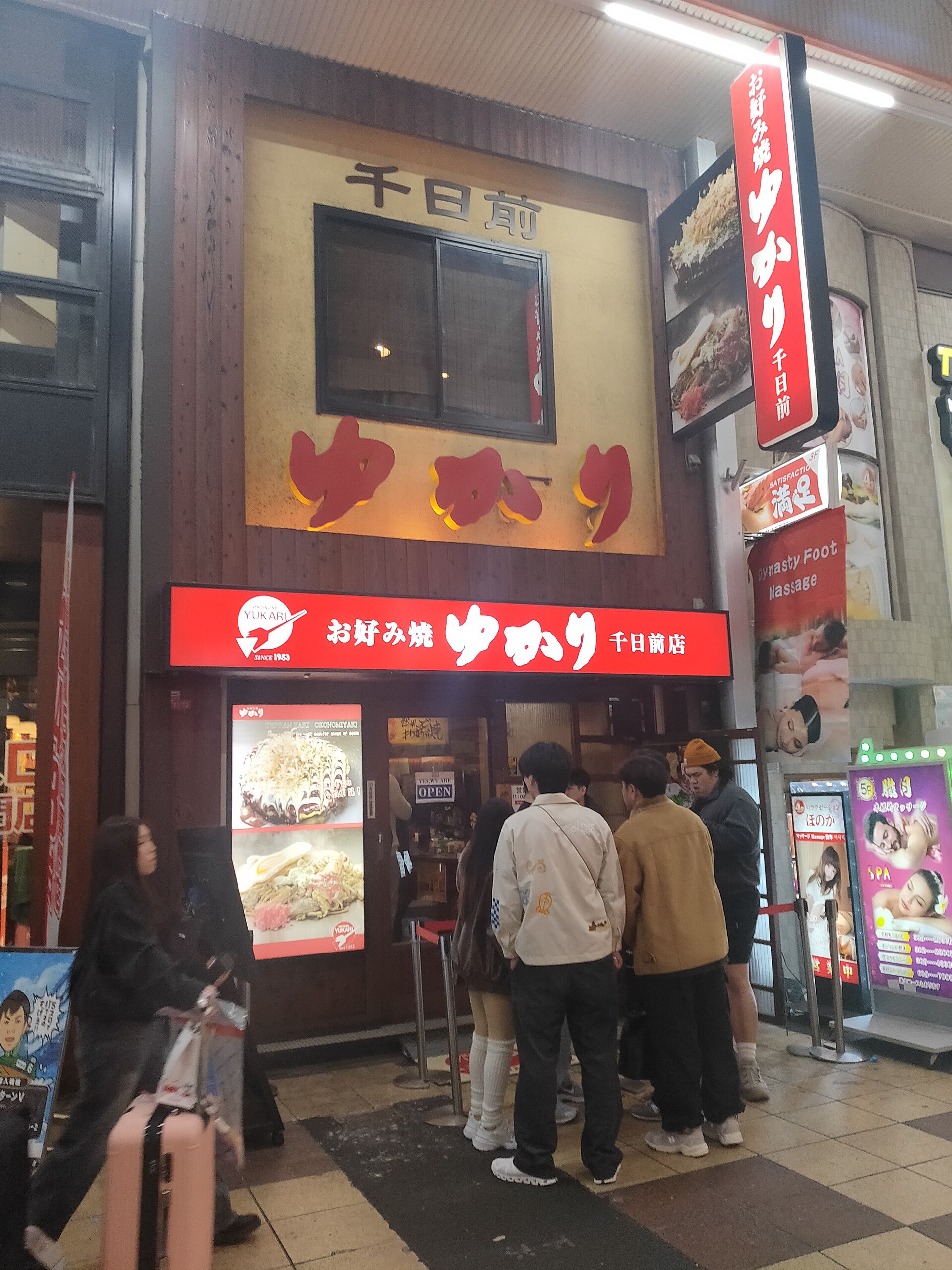 Customers stand at Yukari okonomiyaki restaurant beneath red illuminated signs in the Sennichimae area of Osaka.