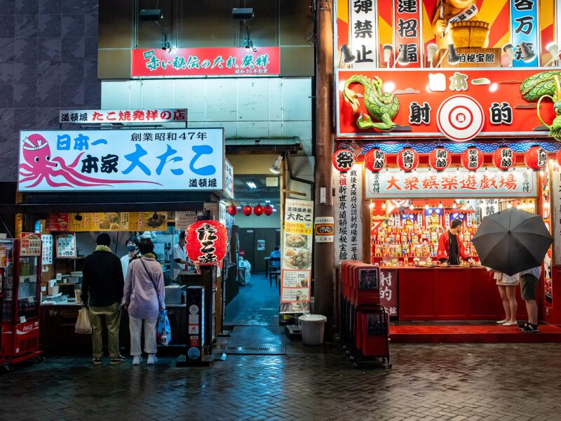 Customers visit a brightly lit takoyaki counter in Osaka's Dotonbori at night, beside a red games stall on wet paving.
