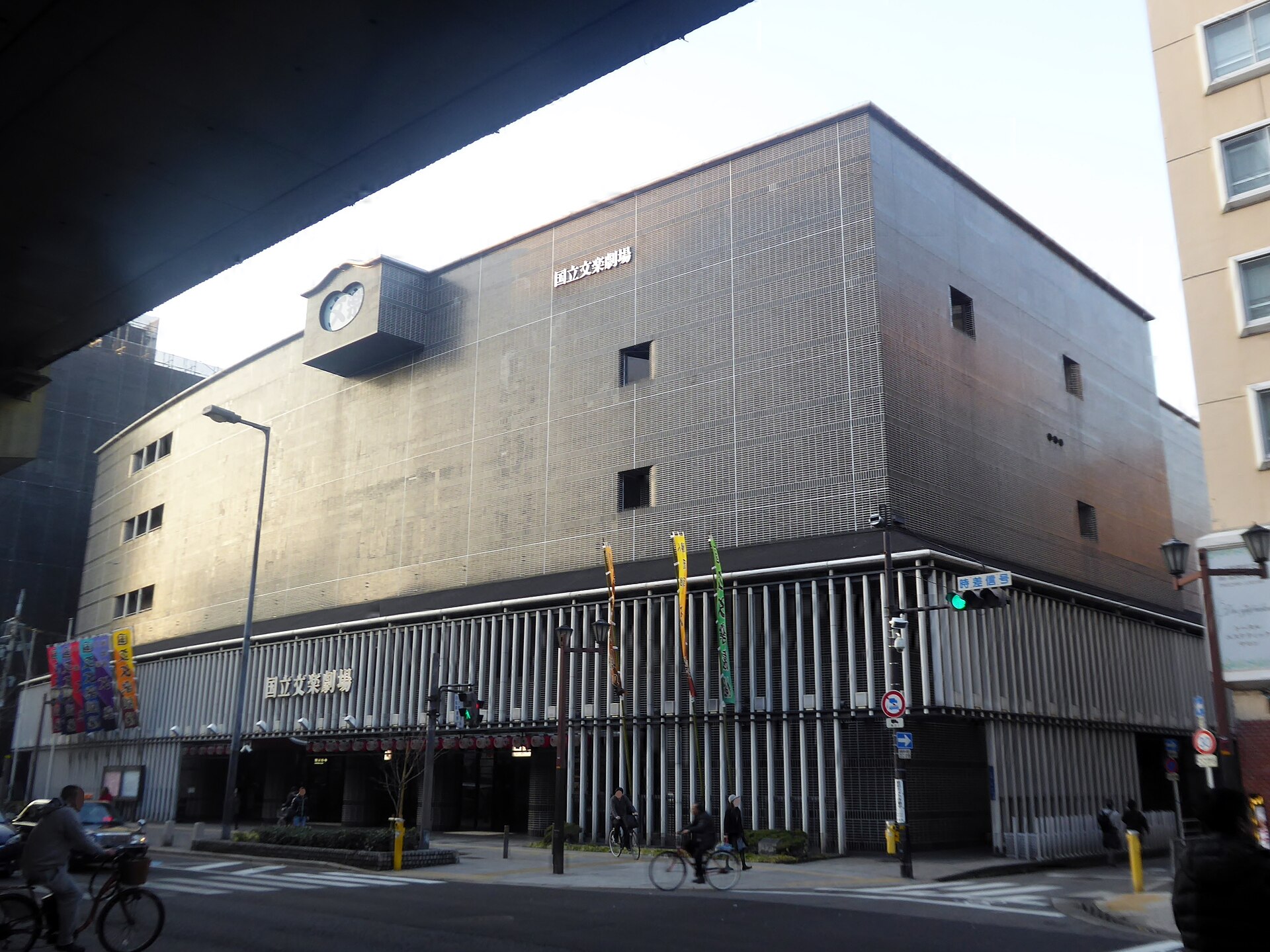Cyclists pass the National Bunraku Theatre in Osaka, beneath its projecting clock and tall grey facade.