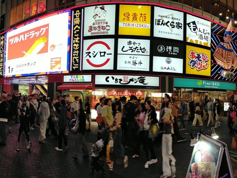 Evening crowds pass the brightly lit Nakaza Kuidaore building in Osaka's Dotonbori, beneath restaurant signs and a striped drummer figure.