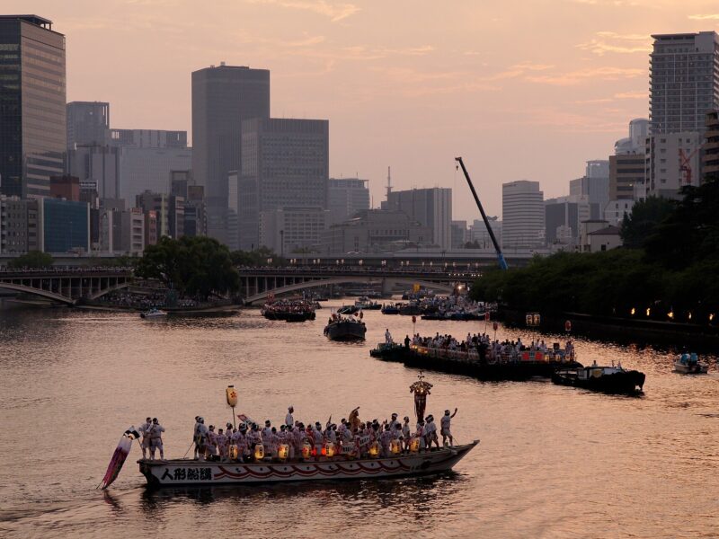 Festival boats fill the Okawa River beneath Osaka's buildings and a pink evening sky during the summer Tenjin Matsuri.