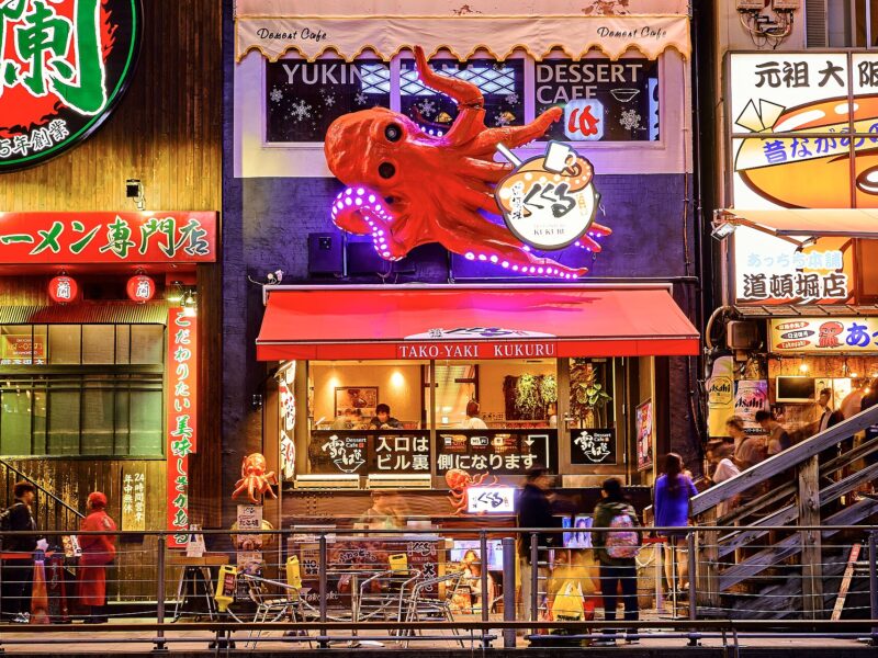A giant red octopus crowns Takoyaki Kukuru in Osaka's Dotonbori, above an illuminated counter and pedestrians on the walkway.
