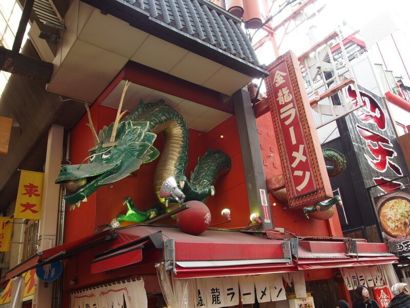 A green dragon sculpture curls above the red entrance of Kinryu Ramen in Dotonbori, Osaka, beside a vertical restaurant sign.