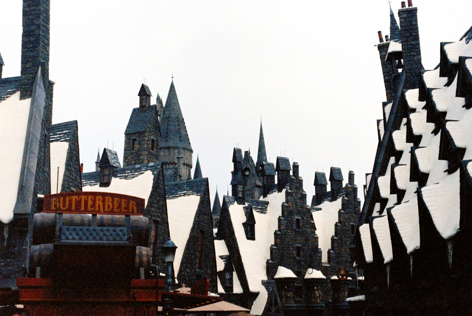 Snow-capped rooftops, crooked chimneys and turrets of a Hogsmeade-style village street, with a red Butterbeer cart sign in the foreground