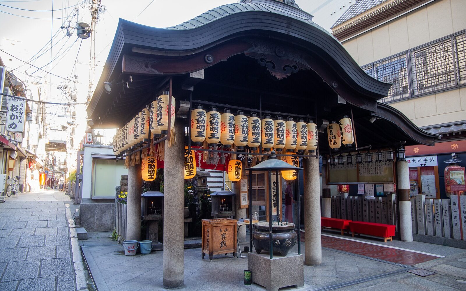Hozenji’s curved-roof worship hall in Osaka stands beside a paved alley, with hanging lanterns, stone columns, water buckets, and an incense vessel.
