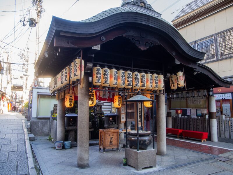 Hozenji’s curved-roof worship hall in Osaka stands beside a paved alley, with hanging lanterns, stone columns, water buckets, and an incense vessel.