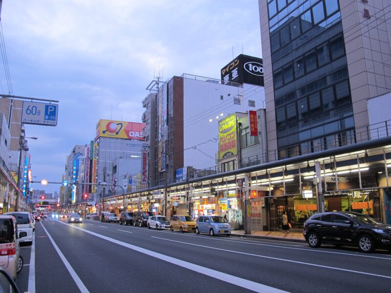 Illuminated electronics shops line a covered pavement beside evening traffic in Osaka’s Den Den Town.