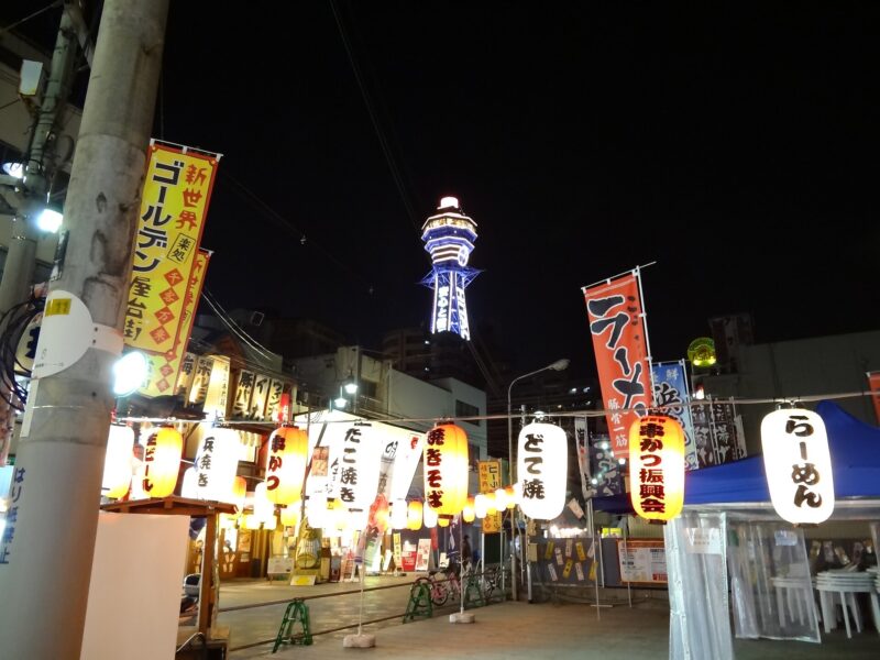 Illuminated Tsutenkaku rises above restaurant signs and glowing paper lanterns along a Shinsekai street in Osaka at night.