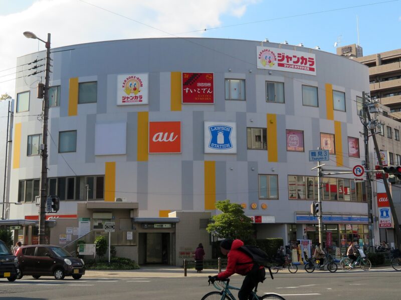 Jankara karaoke signs cover the upper floor of a curved Osaka building as cyclists pass the Lawson store below.