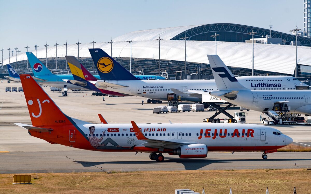 A Jeju Air jet crosses the apron at Kansai International Airport, with parked aircraft and Terminal 1's sweeping silver roof behind.