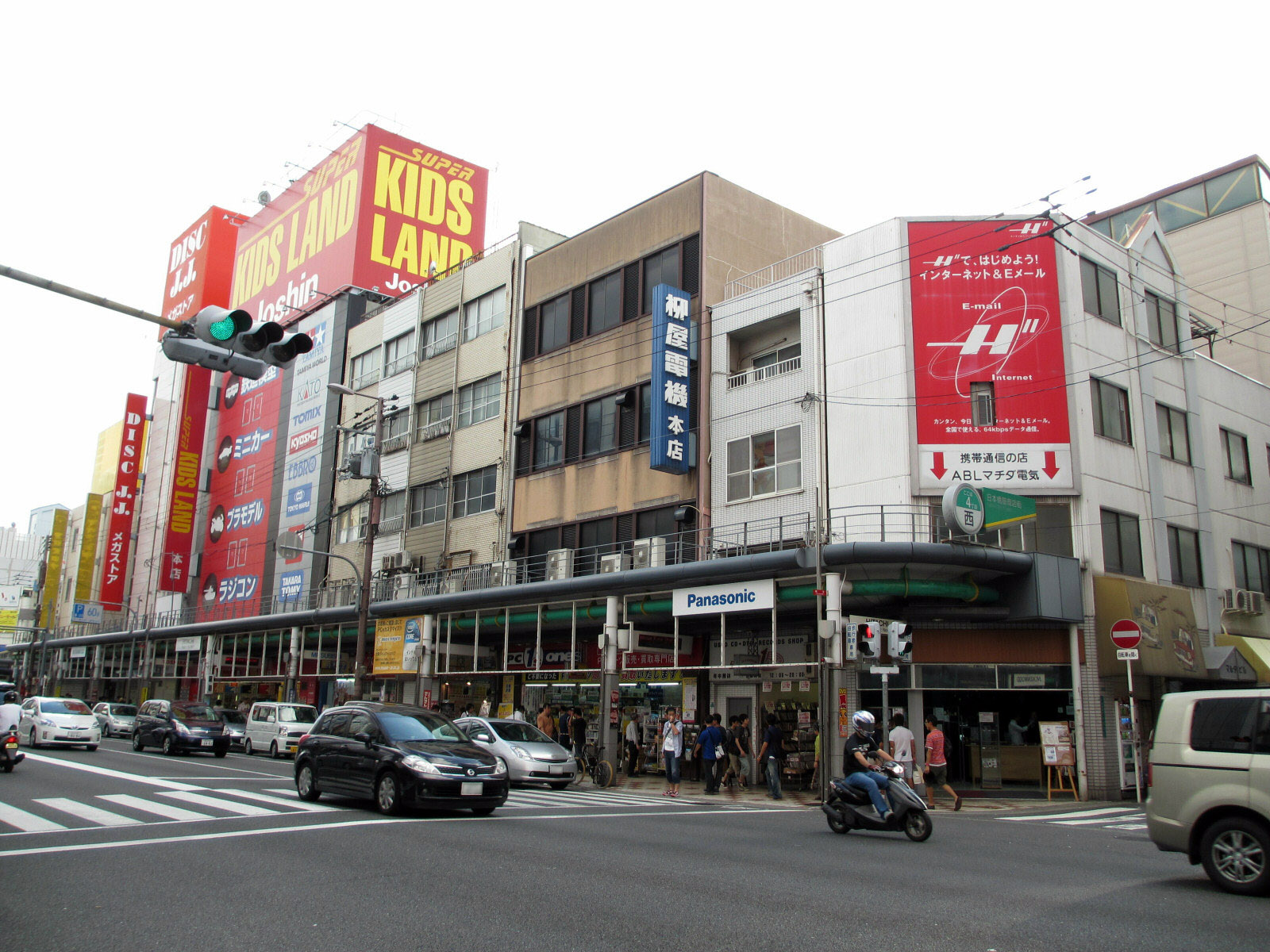 Joshin Kids Land and electronics shop signs rise above covered pavements and traffic in Osaka’s Den Den Town.