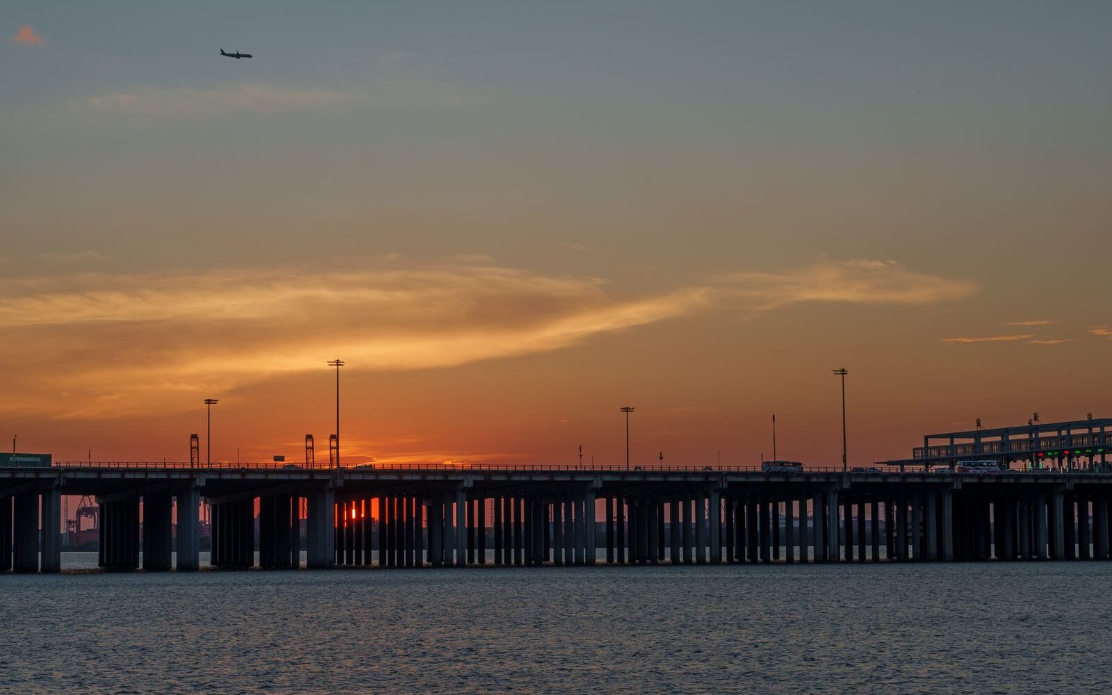 A long double-deck bridge stretching across calm water at sunset, with a plane in the sky above