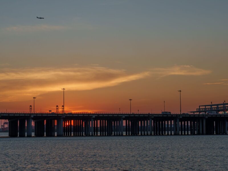 A long double-deck bridge stretching across calm water at sunset, with a plane in the sky above