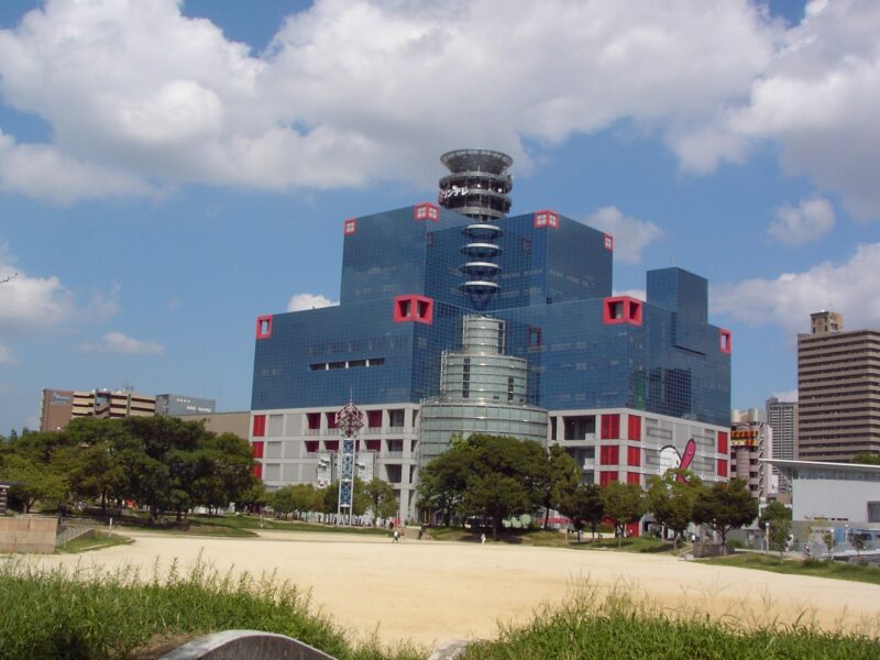 Kids Plaza Osaka occupies a stepped blue-glass building with red window frames beside the open grounds of Ogimachi Park.