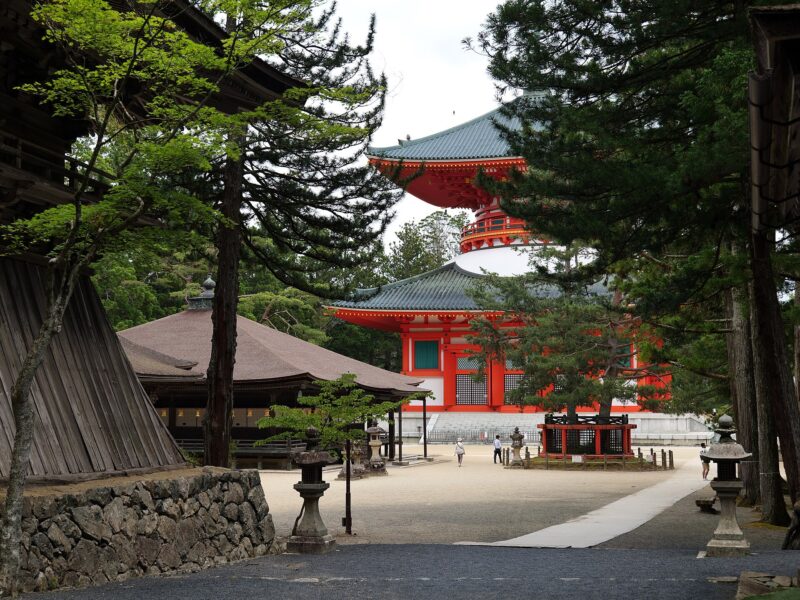 Koyasan's vermilion Konpon Daito pagoda rises behind wooden temple halls and trees, with visitors crossing the gravel courtyard.