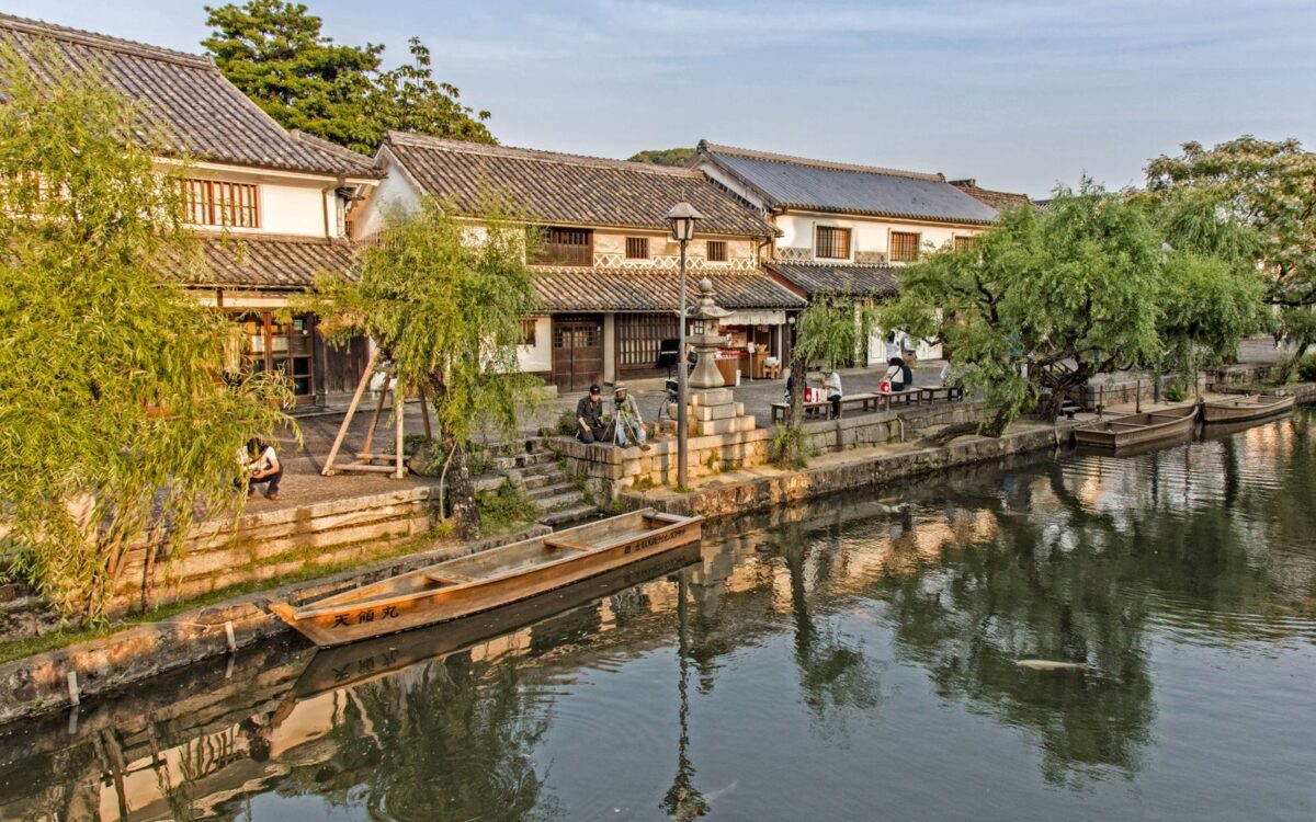 The Kurashiki canal in the Bikan Historical Quarter, lined with willows and traditional tiled buildings, with a wooden boat moored at the bank