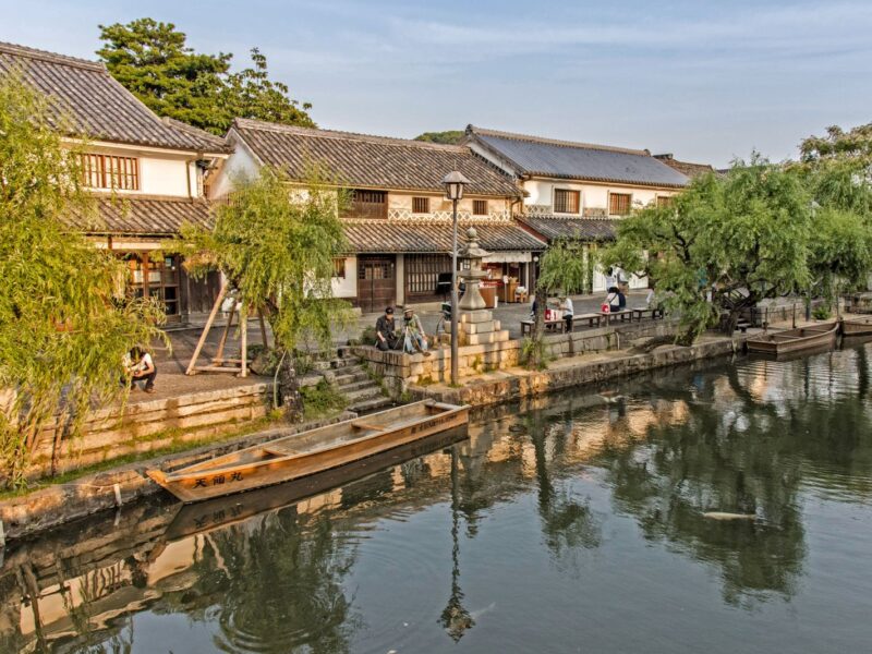 The Kurashiki canal in the Bikan Historical Quarter, lined with willows and traditional tiled buildings, with a wooden boat moored at the bank