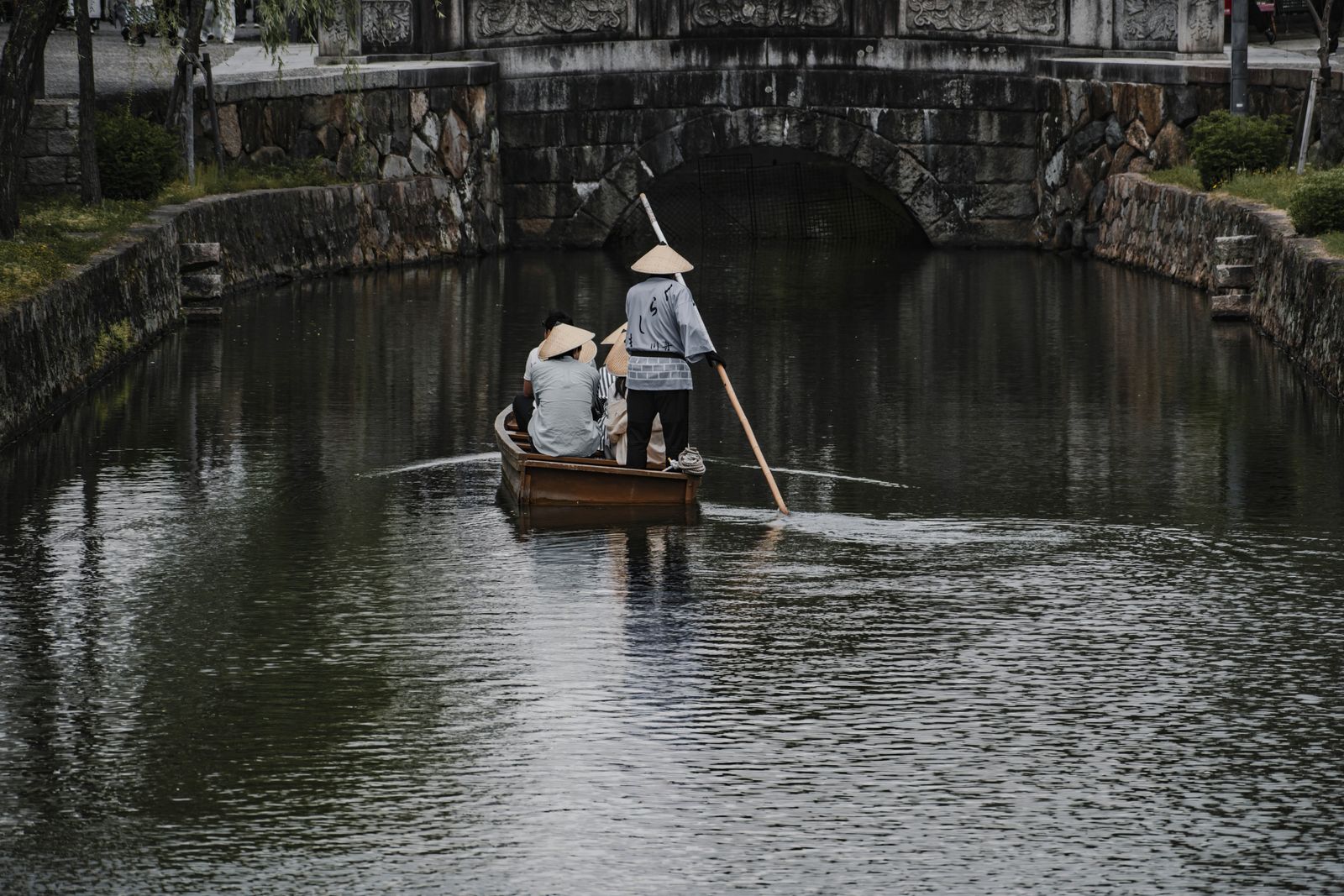 A boatman in a conical sedge hat poling a small wooden boat with passengers along the Kurashiki canal towards a stone bridge
