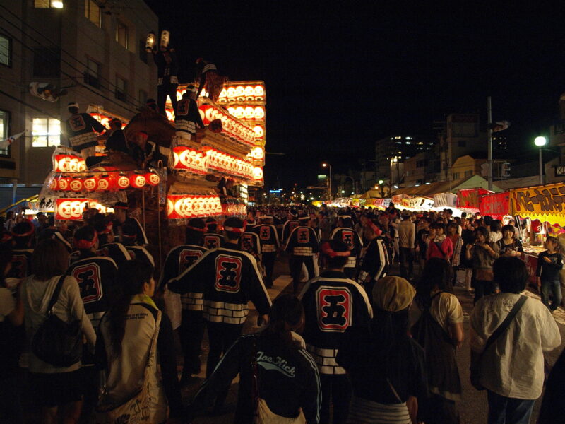 Lantern-covered floats glow above a crowd on a stall-lined street during Kishiwada Danjiri Matsuri in Osaka Prefecture at night.